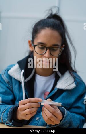 Pretty young female in casual outfit rolling cigarette while sitting on street Stock Photo