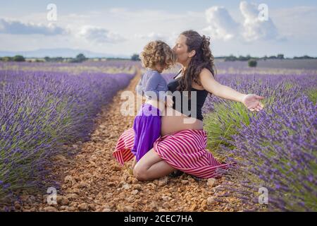 Side view of happy pregnant Woman kissing little daughter while sitting on pathway in blooming lavender field Stock Photo