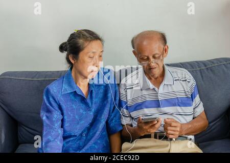 Asian elderly couples watch mobile phones and use headphones on the sofa. Stock Photo