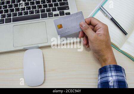 Top view man holding a credit card and wearing a blue plaid shirt on the desk. Laptop computer Notepad and pen.Online shopping business, pay by credit Stock Photo