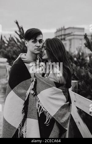 Beautiful young couple stand face to face among christmas tree. Young newlyweds in love, gently kissing, hugging, dressed in warm clothes, snow-covered city background, love story. Black and white photo. Stock Photo