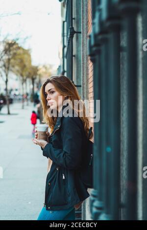 Side view of pretty Woman standing with paper cup of coffee on street. Stock Photo