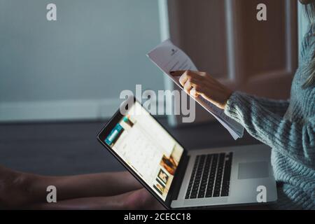 Crop unrecognizable Woman sitting on floor and reading papers at laptop. Stock Photo