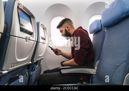 Side view of handsome bearded male browsing smartphone while sitting on comfortable seat inside modern aircraft Stock Photo