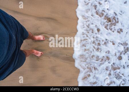Crop shot from above of barefoot man standing on smooth sandy beach with running foamy wave Stock Photo