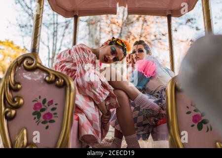 Two pretty young women with theatrical makeup and costumes looking at camera while sitting on amazing roundabout in amusement park Stock Photo