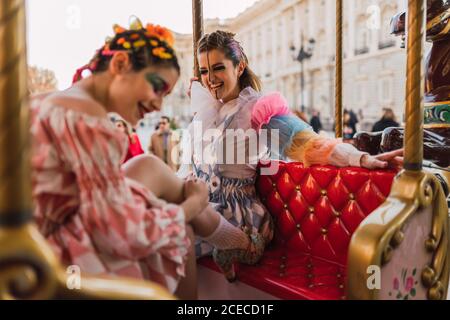 Two pretty young women with theatrical makeup and costumes laughing while sitting on amazing roundabout in amusement park Stock Photo
