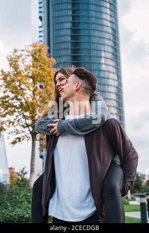 Young man carrying charming girlfriend on back while posing on urban background. Stock Photo