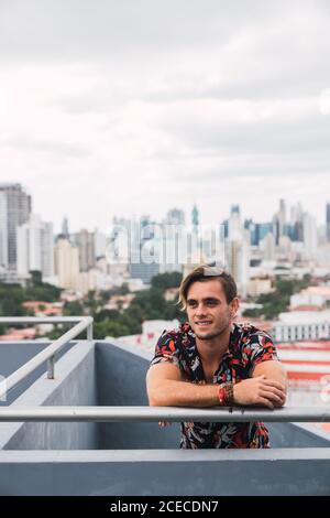Handsome young guy smiling and looking away while leaning on terrace railing on background of modern city on cloudy day Stock Photo