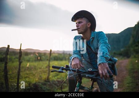 LA HABANA, CUBA - NOVEMBER, 6 , 2018: Young man in jean jacket and hat smoking cigar and cycling on countryside road in Cuba Stock Photo