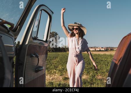 View from inside of retro car of happy trendy Woman in hat and sunglasses standing excitedly in nature and laughing Stock Photo