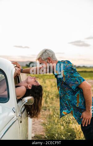 Side view of Woman sitting inside of car leaning head out and looking at man standing above and smiling happily in summertime Stock Photo