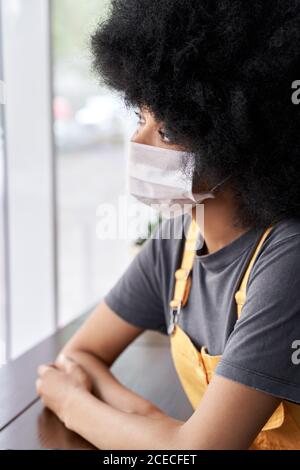 Thoughtful pensive African lady wearing face mask looking through window. Stock Photo
