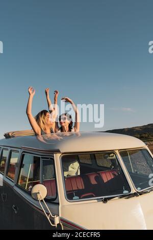 Two joyful young ladies laughing and holding hands up while standing in hatch in roof of vintage van in beautiful countryside on sunny day Stock Photo