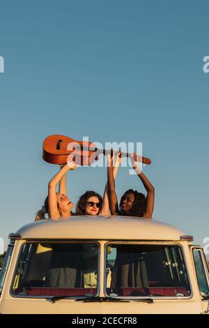 Three cheerful young ladies holding guitar and having fun while standing in hatch in roof of retro van in nature Stock Photo