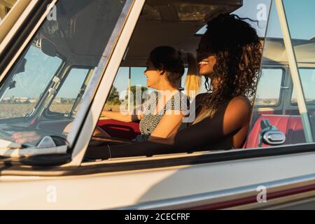 Side view of two young women sitting inside vintage van and smiling while traveling in nature together Stock Photo