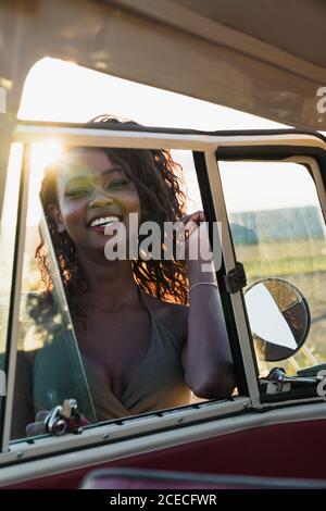Lovely African-American Woman smiling and looking at camera through window of vintage car while spending time in nature on sunny day Stock Photo