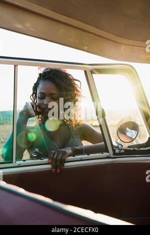 Lovely African-American Woman smiling and looking at camera through window of vintage car while spending time in nature on sunny day Stock Photo