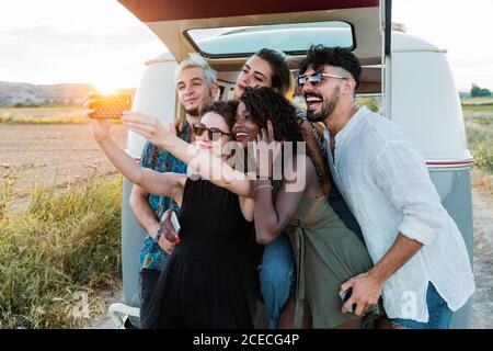 Group of diverse friends smiling and posing for selfie while standing near vintage van in countryside Stock Photo