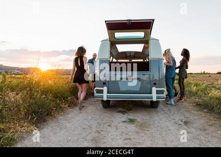 Group of cheerful young people standing near retro van and laughing while spending time in nature together Stock Photo