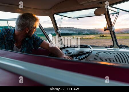 Handsome young man leaning on steering wheel of retro car and looking away while traveling in nature Stock Photo