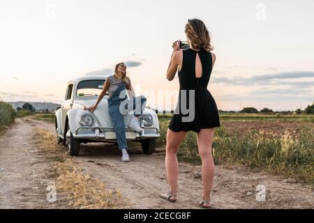 unrecognizable Woman using smartphone to take picture of beautiful Woman leaning in a old car in nature Stock Photo