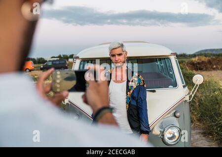 unrecognizable man using smartphone to take picture of handsome young guy standing near van in nature Stock Photo