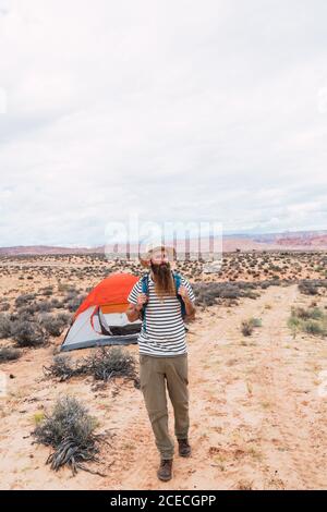 Handsome bearded man with backpack looking away while walking near tent on cloudy day in desert Stock Photo