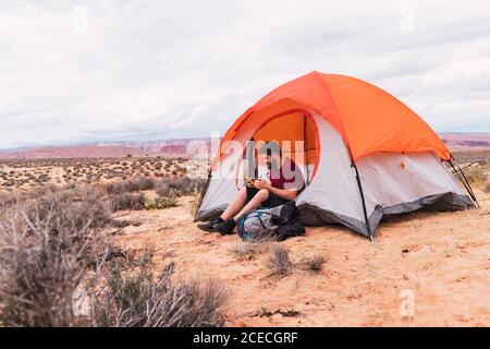 Bearded guy in casual outfit holding mug of hot beverage and modern smartphone while sitting near tent and looking away in beautiful nature Stock Photo