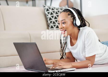 Excited beautiful young Black woman in dreadlocks lying on the floor and studying or working on laptop Stock Photo