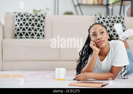Portrait of pensive beautiful young Black woman lying on the floor with cup of coffee and digital tablet Stock Photo