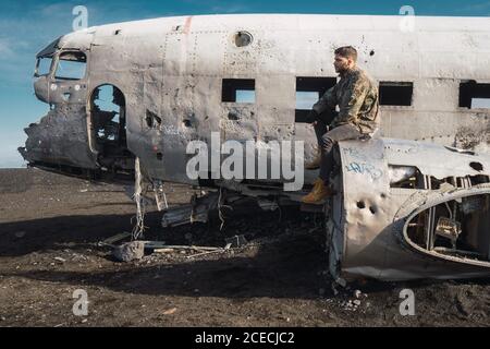 Side view of young male sitting on old crashed aircraft outdoors and looking away Stock Photo