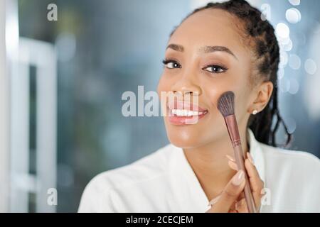 Beautiful attractive young Black woman applying peach blush on her cheeks when getting ready in the morning Stock Photo