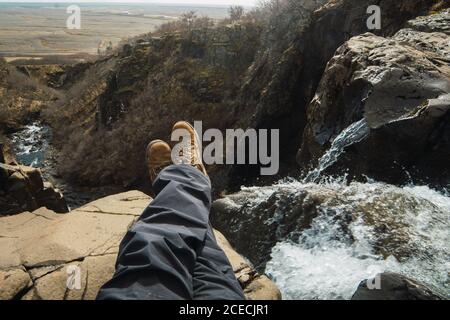 Crop legs of male sitting on top of mountain near waterfall in winter clothes Stock Photo