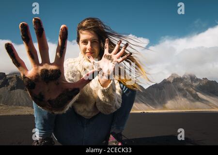 Beautiful Woman with waving hair showing hands in black sand smiling at camera on background of mountains, Iceland. Stock Photo