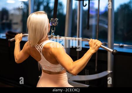 Back view of attractive young Woman in sportswear doing Lat Pull Down exercise on gym equipment near window Stock Photo