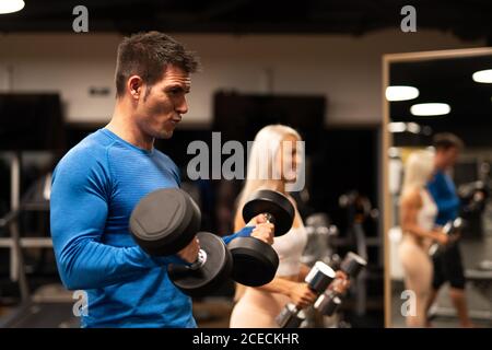 Side view of handsome man in blue pullover and attractive Woman in sportswear standing in gym in front of mirror and doing exercise with dumbbells Stock Photo