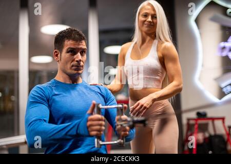 Young handsome man in blue pullover doing Seated Cable Raw exercise in gym with pretty blond Woman standing near and looking at camera Stock Photo