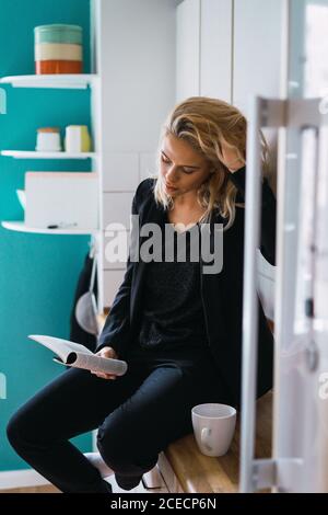 Casual relaxed Woman having coffee while sitting on kitchen counter in morning reading book. Stock Photo