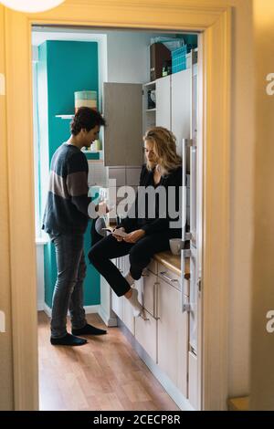 Young casual couple spending time together in kitchen reading and having drink. Stock Photo