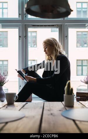 Young casual model immersed in book while lounging on window sill of apartment. Stock Photo