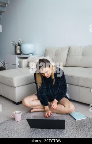 Casual blond Woman using laptop while sitting on floor at home having coffee cup near. Stock Photo