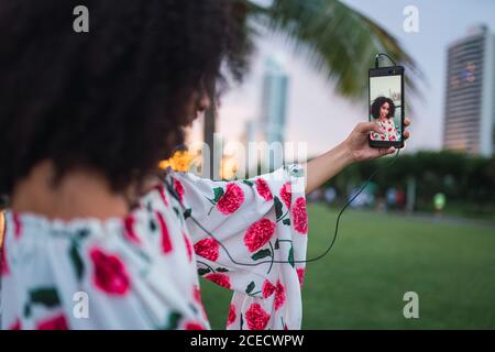 Cheerful black Woman using phone taking selfie while listening to music on street of Panama city in twilight Stock Photo