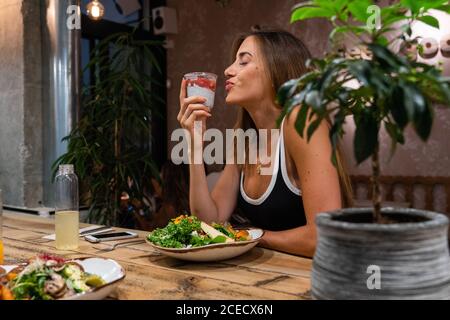 Side view of beautiful young female sitting at wooden table with delicious food and holding plastic cup full of sweet tasty dessert with closed eyes a Stock Photo