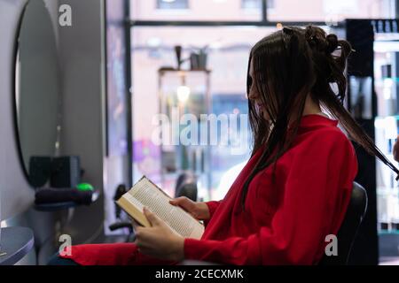 Side view of attractive young lady reading volume and sitting on chair with beautiful hairstyle in hairdressing salon Stock Photo
