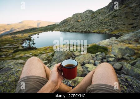 Crop man with metal mug having warm tea while sitting on rocky coast of small lake in mountains, Spain Stock Photo