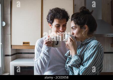 Happy young couple standing with two cups and smiling on kitchen in the morning. Stock Photo