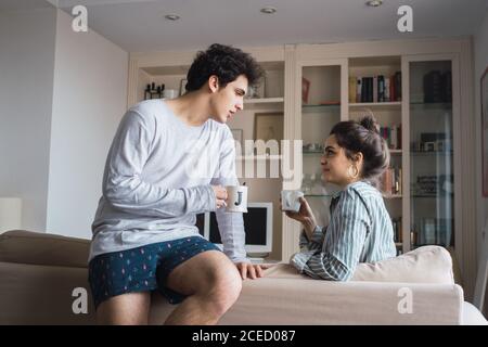 Happy young couple in pajamas sitting on sofa in living room with cups. Stock Photo