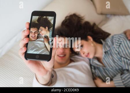 From above cheerful young man and Woman lying on bed and taking selfie with smartphone. Stock Photo