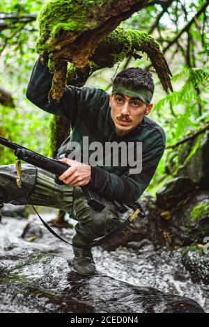 Soldier or revolutionary member or hunter in camouflage in the river observing the gun in his hand Stock Photo